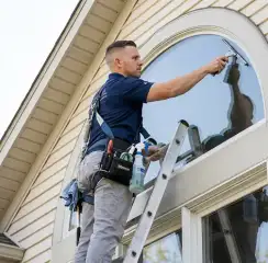 Professional window cleaner on a ladder using a squeegee to clean an exterior arched window, Aurora, CO.